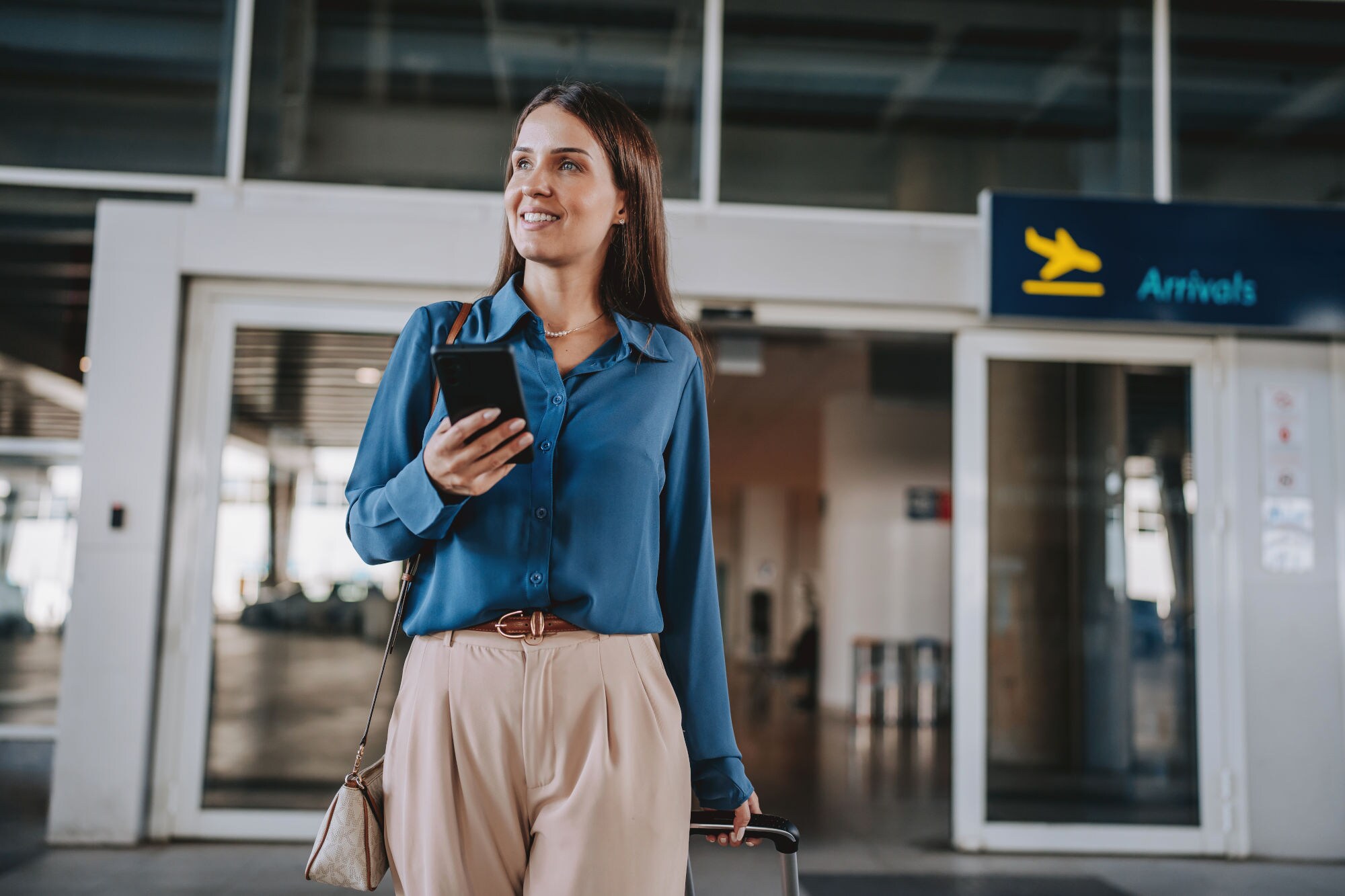 Eine elegant gekleidete Frau mit Smartphone steht vor dem Eingang der Ankunftshalle eines Flughafen-Terminals.